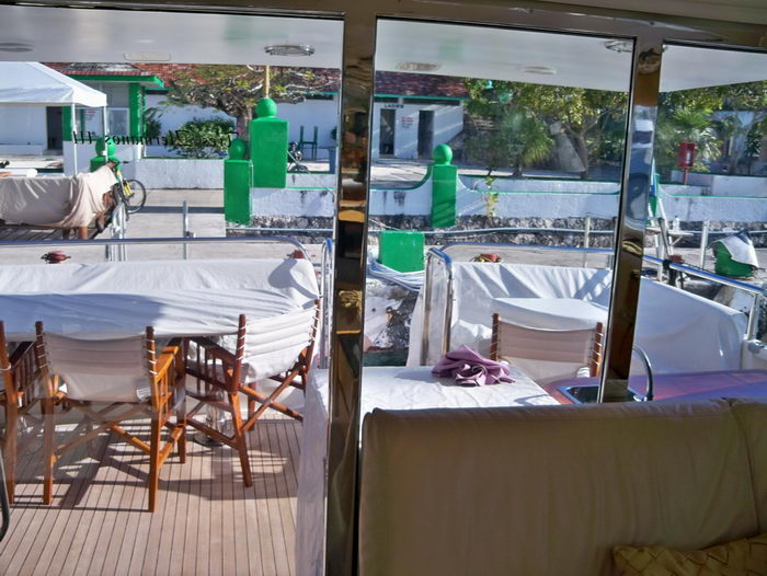 View from inside a yacht salon through glass doors out to the shaded aft deck, set with wooden directors' chairs and a draped dining table. A marina, green dock bollards and white buildings are visible beyond; reflections cross the glass.