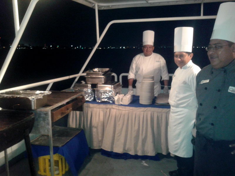 Chefs in white hats at a deck grill and buffet station serving a night dinner cruise on the Discovery with Cancun lights behind