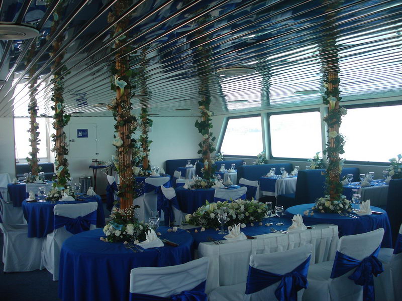 Wedding reception salon on the Discovery boat with round tables in royal-blue and white linens, draped chairs and floral columns by panoramic windows