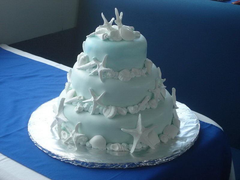 Three-tier seafoam-blue beach wedding cake decorated with white fondant seashells and starfish on a blue-clothed table