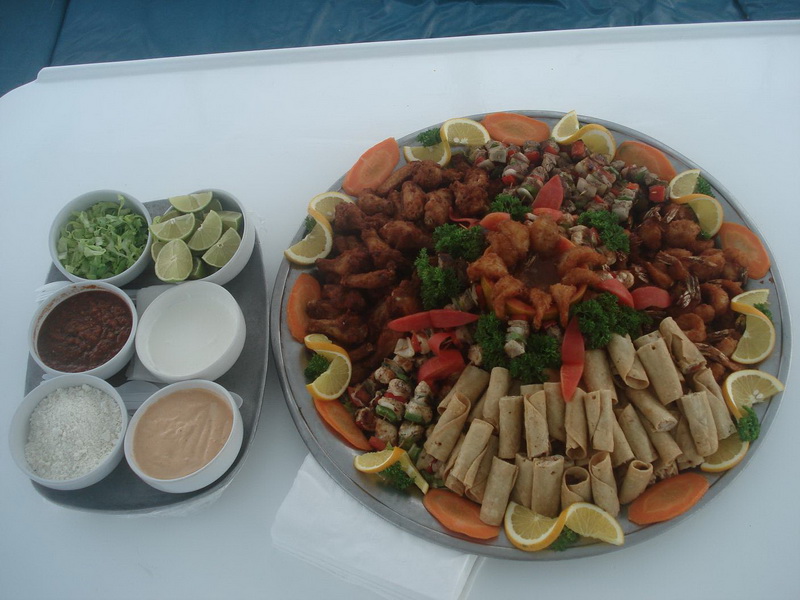 Large round platter of fried appetizers, taquitos, kebabs and vegetable garnish beside a tray of dipping sauces and lime wedges, set on a white table aboard the catamaran.