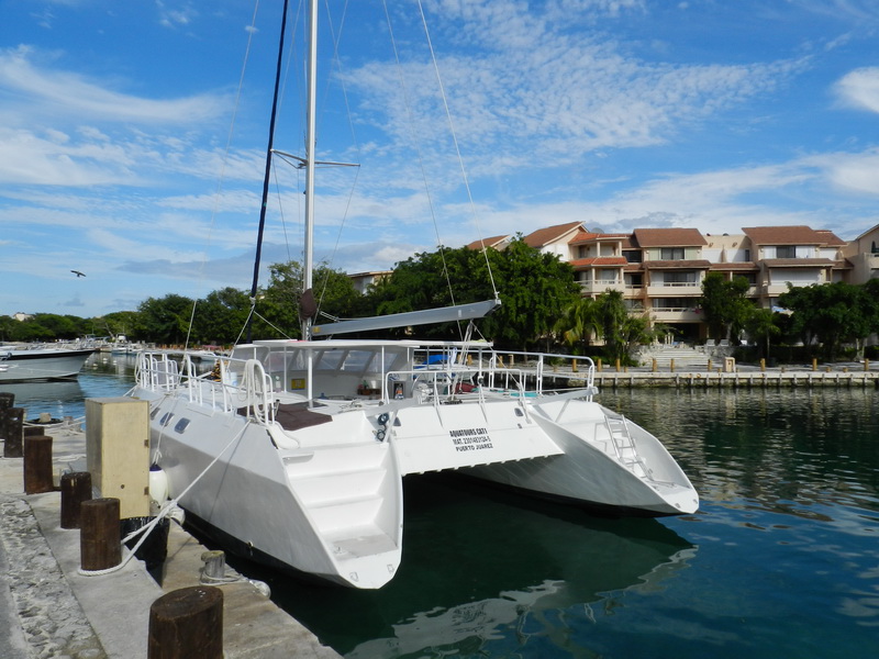 White sailing catamaran with tall mast moored bow-in at a marina dock, tied to a concrete quay. Resort condominium buildings and trees behind, other boats to the left, sunny sky with wispy clouds and calm green harbor water.