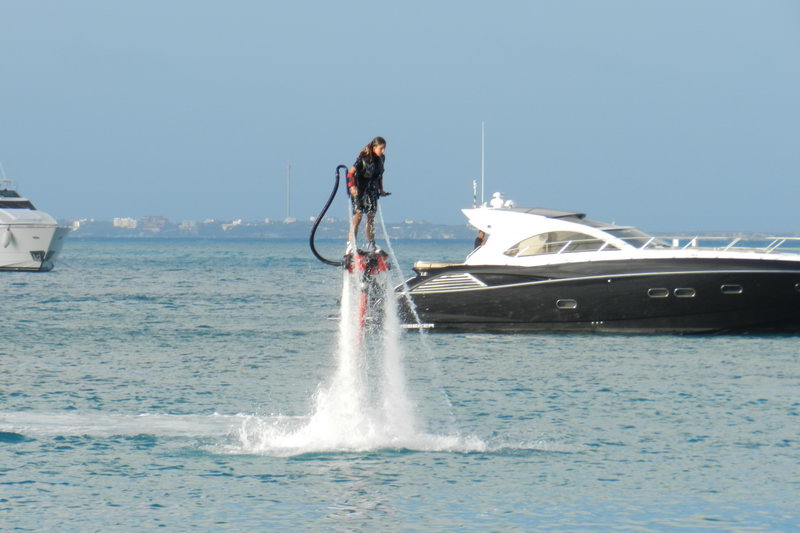 Persona haciendo flyboard suspendida sobre chorros de agua junto a un yate de lujo negro en la tranquila bahía de Cancún con la costa al fondo