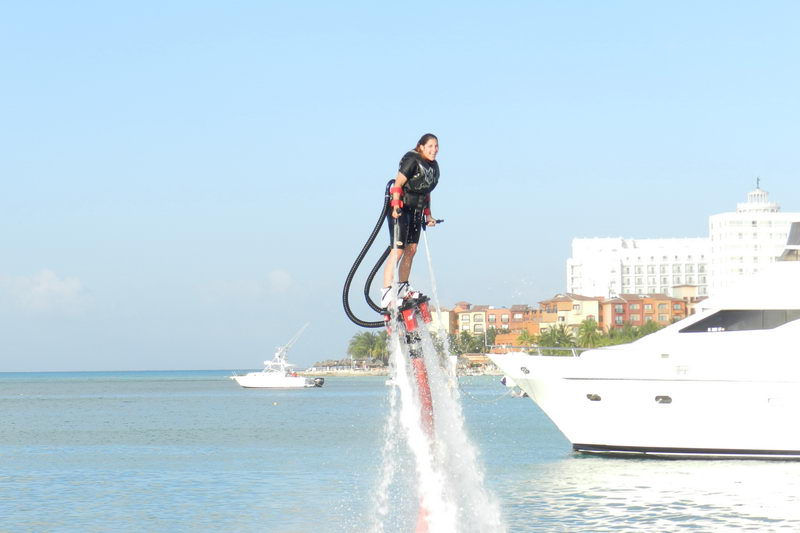 Persona haciendo flyboard elevándose junto a un yate de motor blanco con el skyline de la zona hotelera y la playa de Cancún al fondo