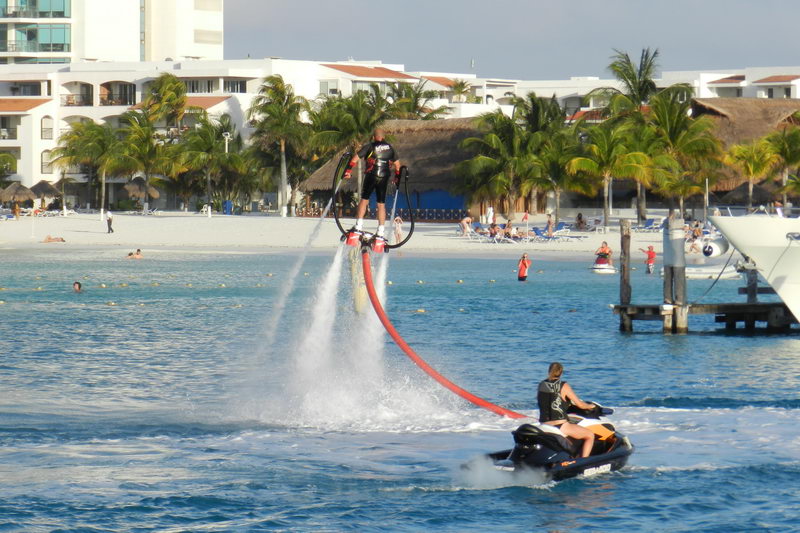 Rider de flyboard suspendido sobre la laguna turquesa frente a un resort de playa de Cancún mientras un instructor observa desde una moto acuática