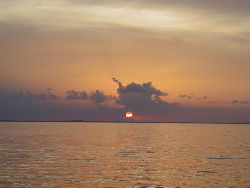 Sunset over a calm bay or lagoon, the sun a red disk sinking behind a low cloud bank near the horizon. Warm orange and grey sky reflected on rippled water, distant land barely visible.