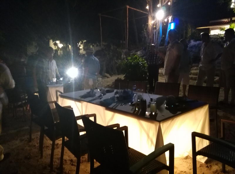 Dark nighttime photo of a beachfront dinner setup, a long white-draped table lit by a bright light with place settings, wicker chairs on sand and several people standing around in low light.