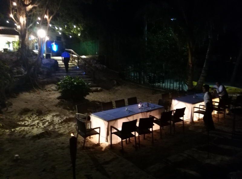 Nighttime beach restaurant scene with two long white-clothed tables and wicker chairs set on sand, lit by string lights and tiki torches, a lit stone staircase and palms behind. Staff barely visible in the dark.