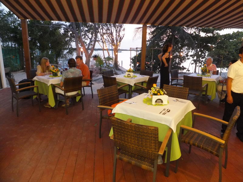 Covered wooden-deck restaurant at dusk with striped awning, set tables in green and white linens, a few diners seated and a waitress standing. Waterfront and palms visible in the fading light behind.