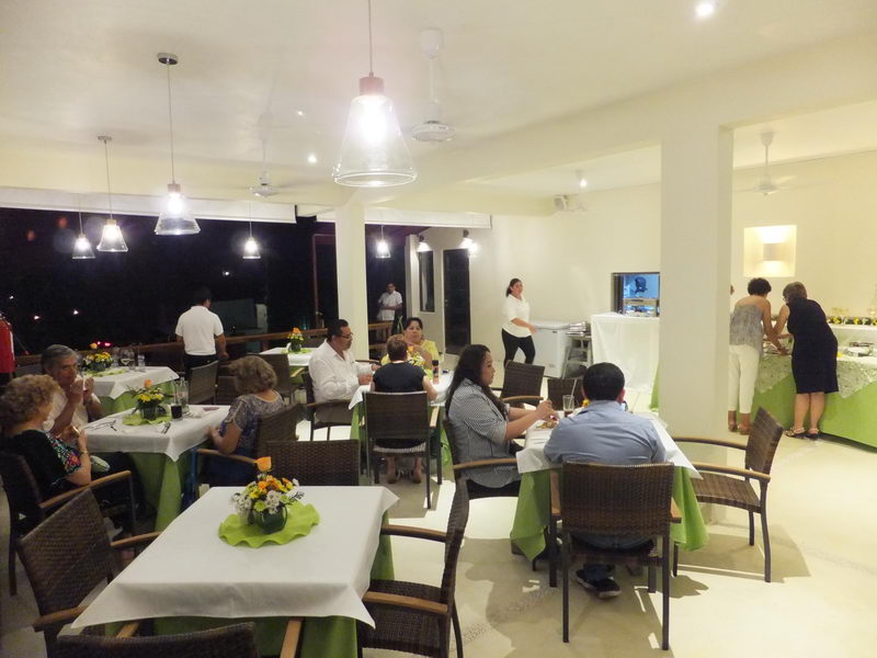 Indoor restaurant dining room at night with white and lime-green table linens, hanging pendant lights and ceiling fans. Numerous guests seated at tables and staff in white shirts serving near a buffet.