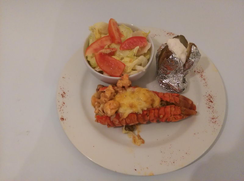 Close-up food photo of a plate with a baked lobster tail topped with melted cheese, a side salad with tomato wedges in a white bowl and a foil-wrapped baked potato with sour cream, dusted with paprika.