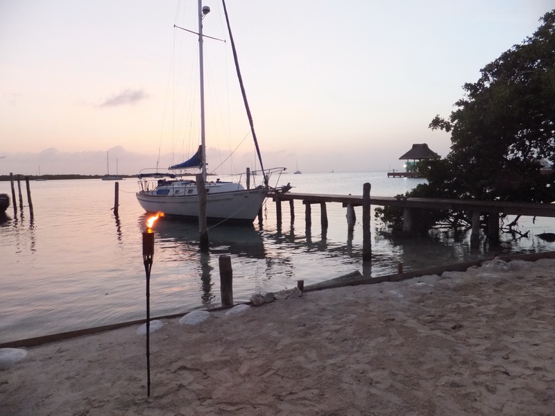 Small white sailboat moored to a rustic wooden dock at dusk, a lit tiki torch in the sandy foreground and a thatched palapa on the water to the right. Soft pink and grey sunset sky over the calm bay.
