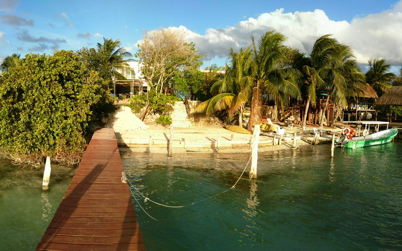 Wooden pier extending from the foreground to a small sandy island with palm trees, buildings and a green boat moored at the far dock. Sunny late-afternoon light, clear water and blue sky with clouds.