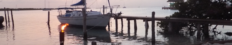 Wide letterbox banner crop of a small white sailboat moored at a wooden dock at dusk with a lit tiki torch in the foreground and a purple-grey sunset sky over the calm bay.