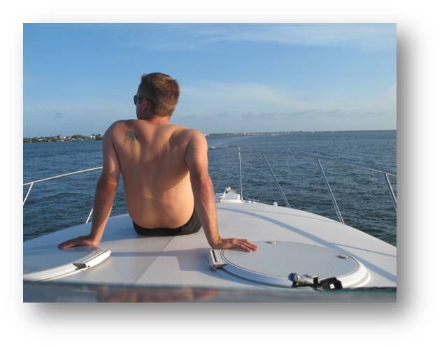 Shirtless man sitting on the white foredeck of a motor yacht with his back to the camera, looking out at a distant coastline and calm sea. Image has a soft grey drop-shadow frame border.