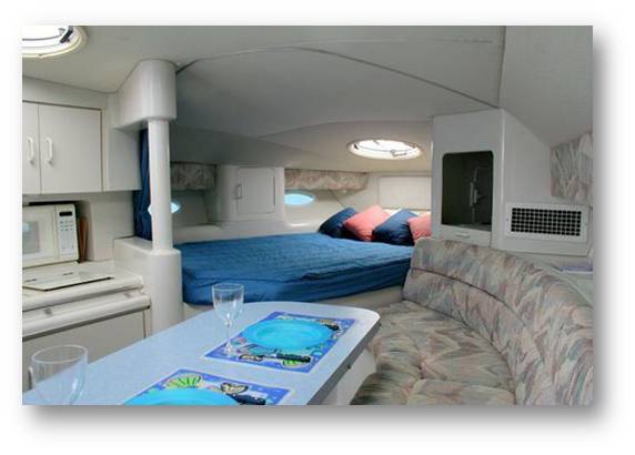 Interior cabin of a motor yacht showing a double bed under portholes, a compact galley with microwave and cabinets, a curved sofa and a table set with wine glasses and themed placemats. Soft grey frame border.
