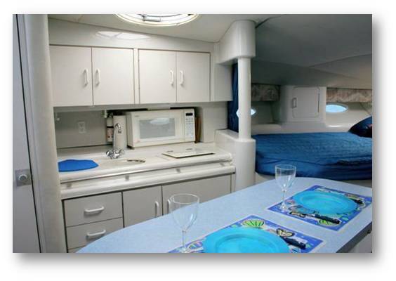 Interior yacht galley and cabin with white cabinetry, a microwave and sink counter, a dinette table set with two wine glasses and placemats, and a blue-covered double berth beyond under a porthole. Grey frame border.