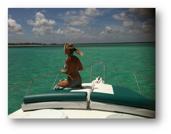 Woman in a bikini kneeling on the green sunpad at the bow of a motor yacht, looking out over brilliant turquoise water toward a low sandy island. Blue sky with clouds; image has a grey frame border.