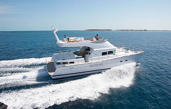 White power catamaran cruising at speed on calm turquoise water, throwing a light wake, with two people relaxing on the flybridge. A low sandy island sits on the horizon under a clear blue sky.