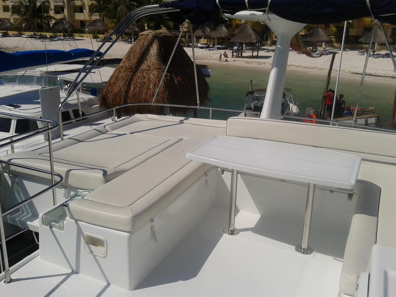 Flybridge sunpad and settee of a catamaran with white cushions and a folding table, overlooking a Cancun beach with thatched palapas, palm trees and clear green water. A crew member in orange vest stands on the dock below. Sunny.