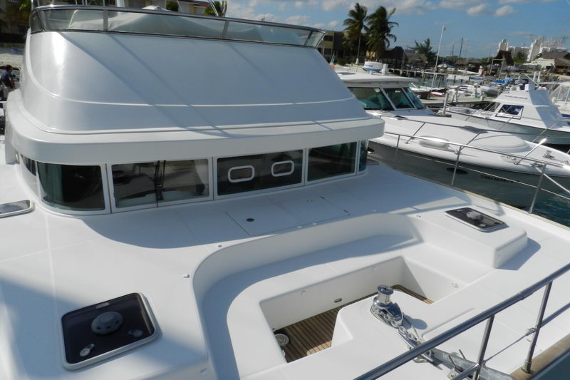 Foredeck and coachroof of a white catamaran with a large windshield and windlass/anchor locker, moored in a marina with several sportfishing boats and palm trees behind. Teak-lined hatch and stainless rails. Sunny.