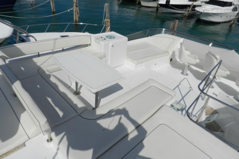 Flybridge lounge of a white motor yacht with a folding table, wraparound white cushioned seating, a pedestal helm seat and console, moored in a marina with other boats behind. Strong midday shadows.