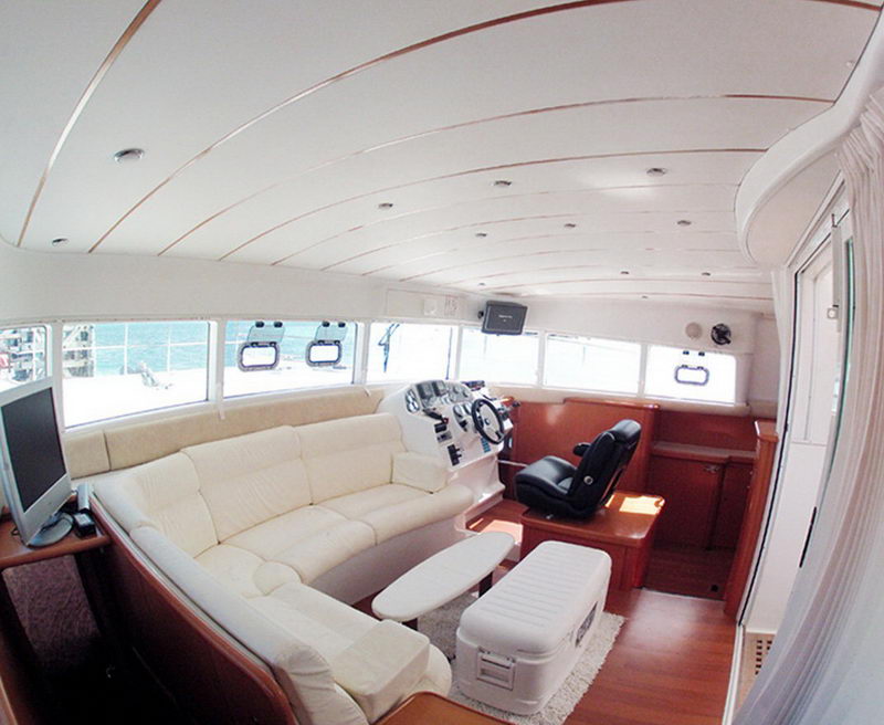Wide-angle interior salon/helm of a catamaran with a cream leather wraparound sofa, wood-trimmed ceiling and cabinetry, an inside steering station with black helm seat, a flat-screen TV and windows looking out to the sea. Bright and airy.
