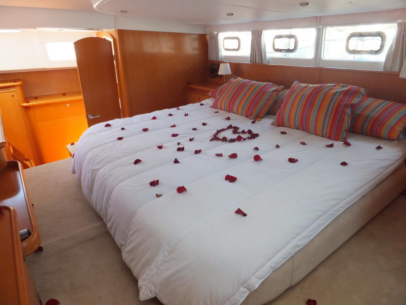 Master cabin of a sailing catamaran with a large double bed made up in white linens scattered with red rose petals arranged in a heart, colorful striped throw pillows, warm wood paneling and small hull windows.