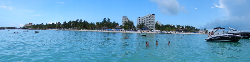 Turquoise Isla Mujeres beach with a motor yacht anchored offshore and swimmers near the palm-lined shore