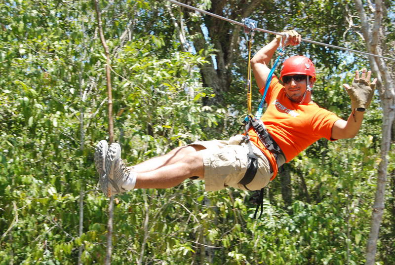 A smiling man in an orange shirt, sunglasses and red safety helmet zip-lines through dense green jungle canopy, arms outstretched, suspended from a steel cable by a harness.
