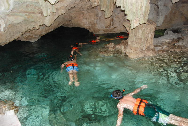 Snorkelers in orange life jackets float single-file through clear turquoise water inside a limestone cavern, surrounded by rock formations and stalactites overhead.