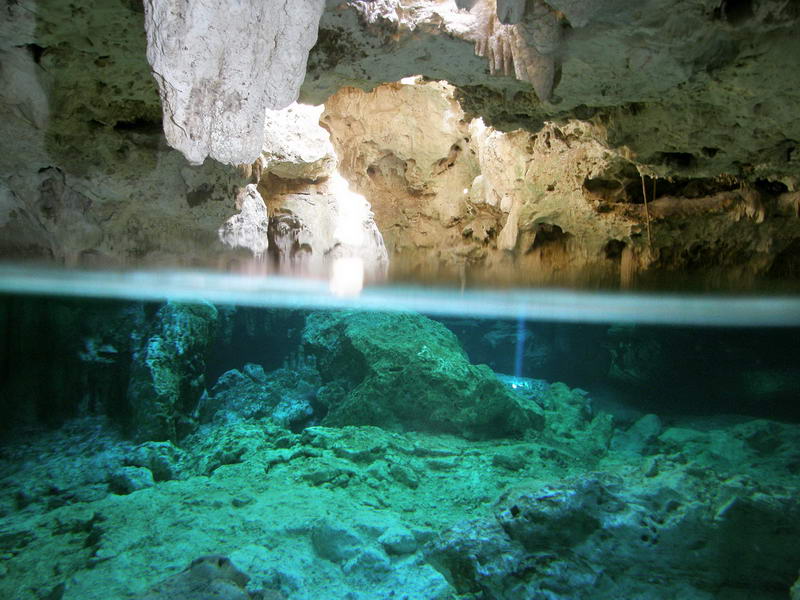 Split above-and-below water view inside a cenote cavern showing rugged limestone rock, hanging stalactites above and clear blue-green submerged rock formations below the waterline.