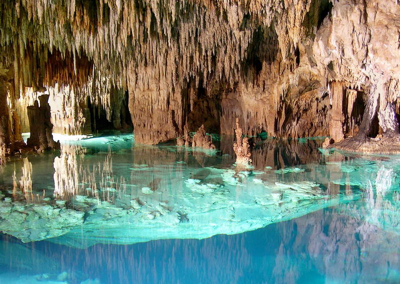 Scenic interior of a limestone cave with dense clusters of stalactites and rock columns mirrored in perfectly still turquoise water. Warm daylight filters in from the left.