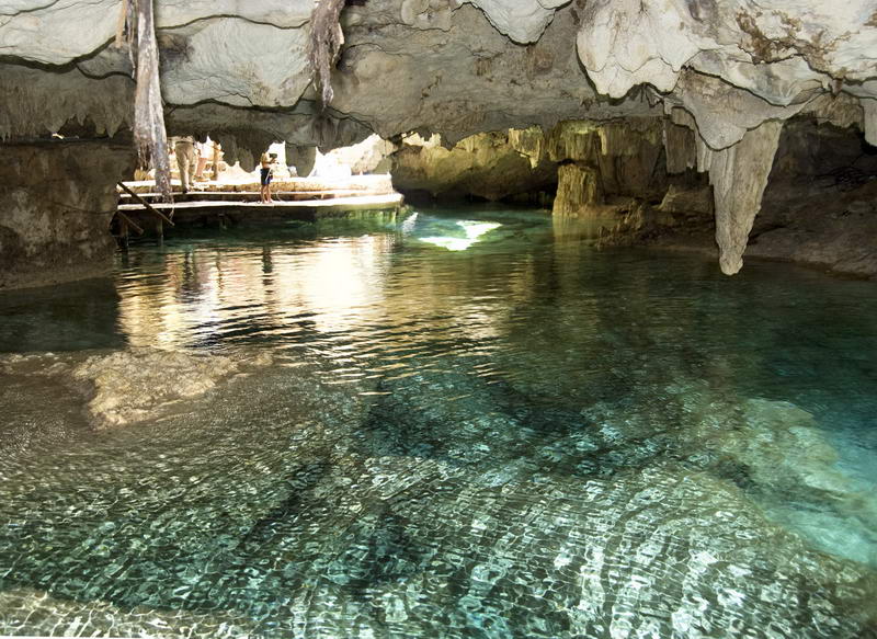 Underground cenote chamber with clear water in the foreground and a wooden viewing platform where small figures stand. Stalactites hang from the ceiling and a shaft of sunlight strikes the water.