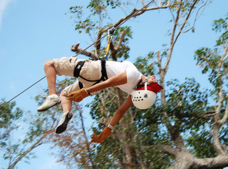 A person zip-lining upside-down/inverted while wearing a white helmet, khaki shorts and gloves, suspended from a cable against a bright blue sky and treetops.