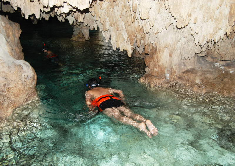 A snorkeler in an orange life jacket floats through clear shallow water inside a dim limestone cavern, with another swimmer visible in the background beneath overhanging rock and stalactites.