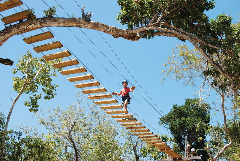 A child in a red shirt and harness crosses a wooden-plank suspension bridge strung high through the treetops against a clear blue sky, part of a jungle adventure course.