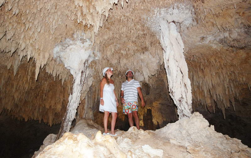 A couple wearing head coverings stand inside a limestone cave densely lined with stalactites and stalagmites, looking upward at the formations.