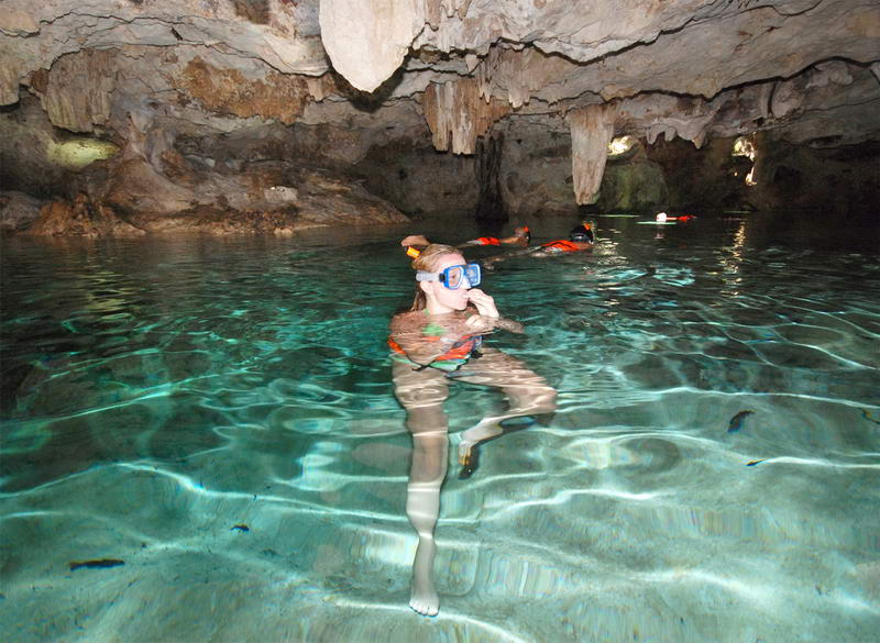 A woman in a blue snorkel mask and orange life jacket floats in crystal-clear cenote water inside a cavern, with other snorkelers behind her under a rock ceiling.
