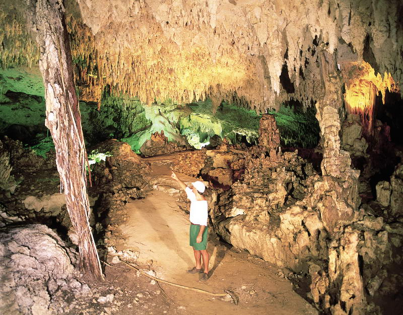 Vast cave interior with dramatic stalactites and stalagmites illuminated in green and amber light. A person in a white shirt stands on the cave floor pointing upward for scale.