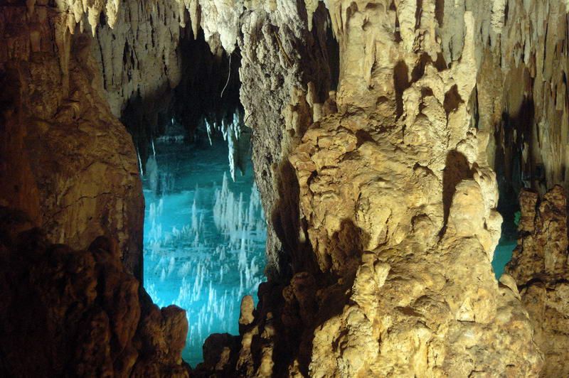 Interior of a limestone cave where towering rock columns and stalactites are reflected in still, brightly-lit blue water in the background.