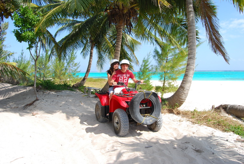 Couple in helmets on a red ATV parked under palm trees beside the white sand and turquoise water of Playa Maroma
