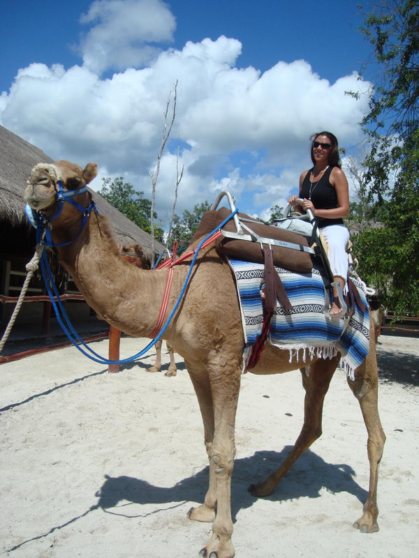Woman seated on a saddled camel beside thatched palapas at the 
