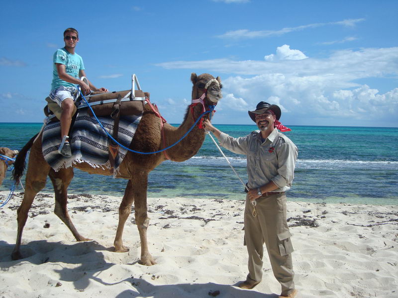 Young man riding a camel led by a smiling guide along the white sand beach beside the turquoise Caribbean at Maroma