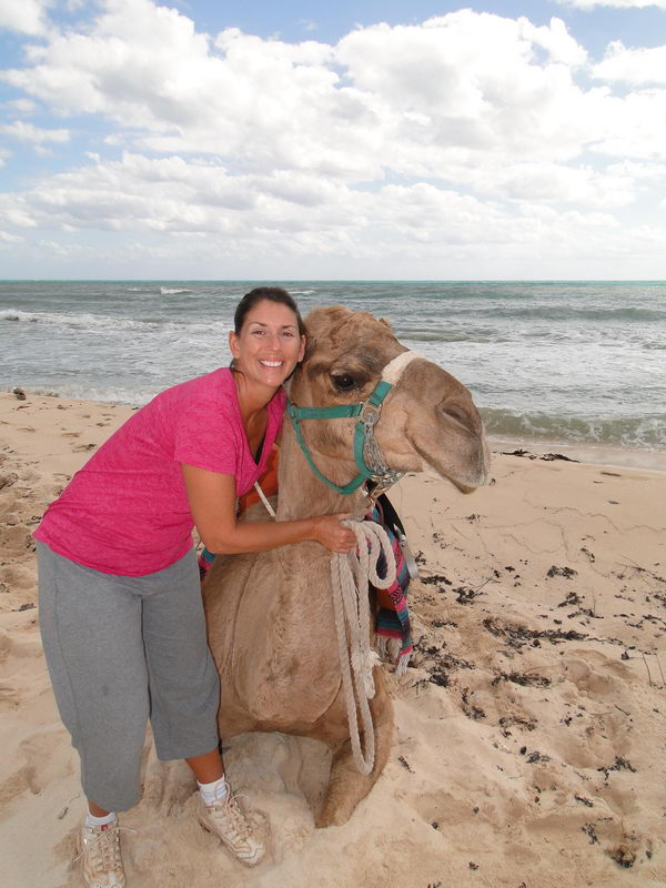 Woman in a pink shirt hugging a resting camel on the sandy Maroma beach with the Caribbean Sea behind