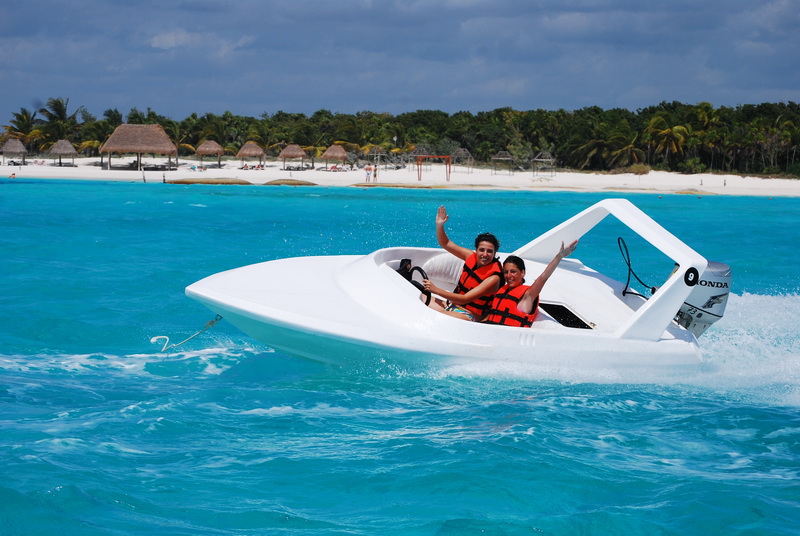 Two people in life vests waving from a small white speed boat skimming the turquoise water off Playa Maroma's white sand beach