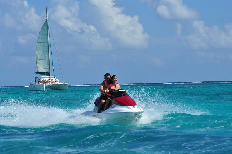 A couple rides a red and white jet ski across bright turquoise water throwing up spray, with a sailing catamaran full of passengers in the background under a blue sky.