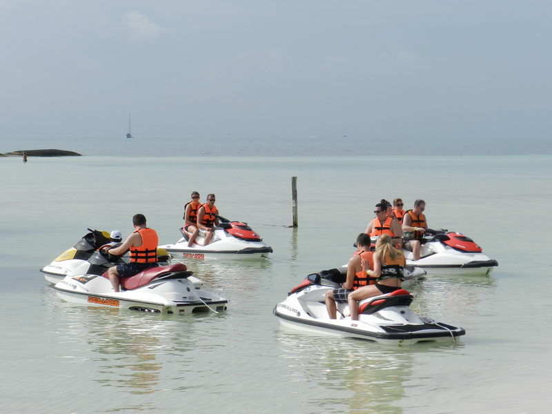 Five personal watercraft (Sea-Doo jet skis, red and white) floating in shallow turquoise water near a beach, ridden by groups of tourists wearing orange life vests. A distant sailboat and hazy sky on the horizon.