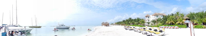 Panoramic beach and marina scene with sailboats moored at left, a motor yacht and small boats offshore, rows of yellow jet skis on the white sand at right, palm trees and beachfront villas in the background under a hazy sky.