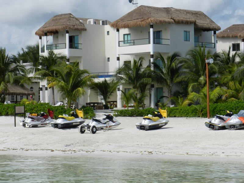 Five personal watercraft (jet skis) parked on white sand in front of a white three-story beachfront villa with thatched palapa roofs and palm trees. Photographed from the water with turquoise sea in the foreground under a cloudy sky.